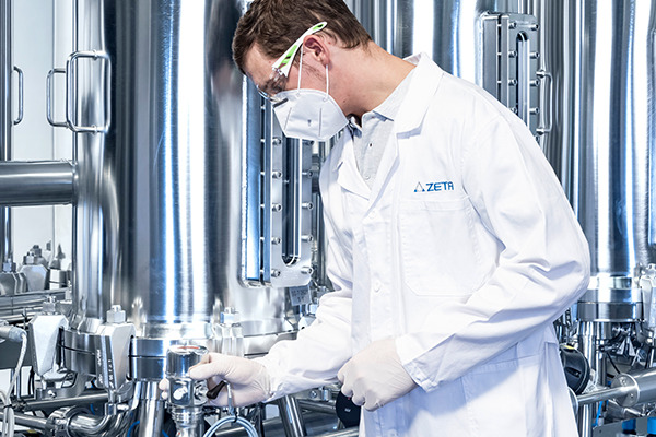 Person with lab coat, face mask and protective gloves handling a device in front of a manufacturing skid