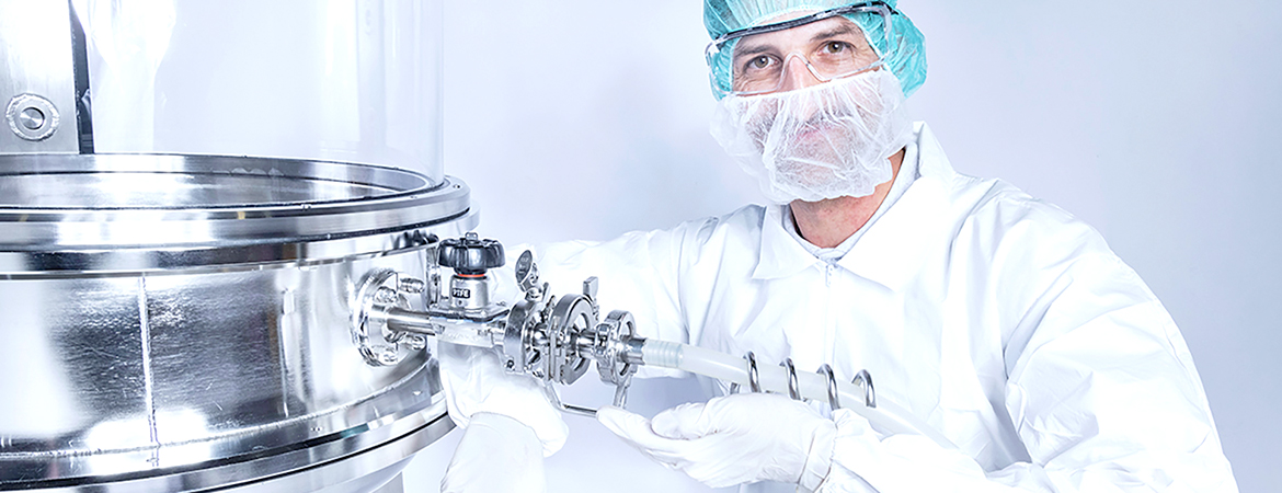 A person dressed in full cleanroom gear—hair cover, face mask, and gloves—is working with a large stainless steel machine. The equipment has several valves and connectors and is likely used in sterile manufacturing, such as pharmaceuticals or biotechnology.