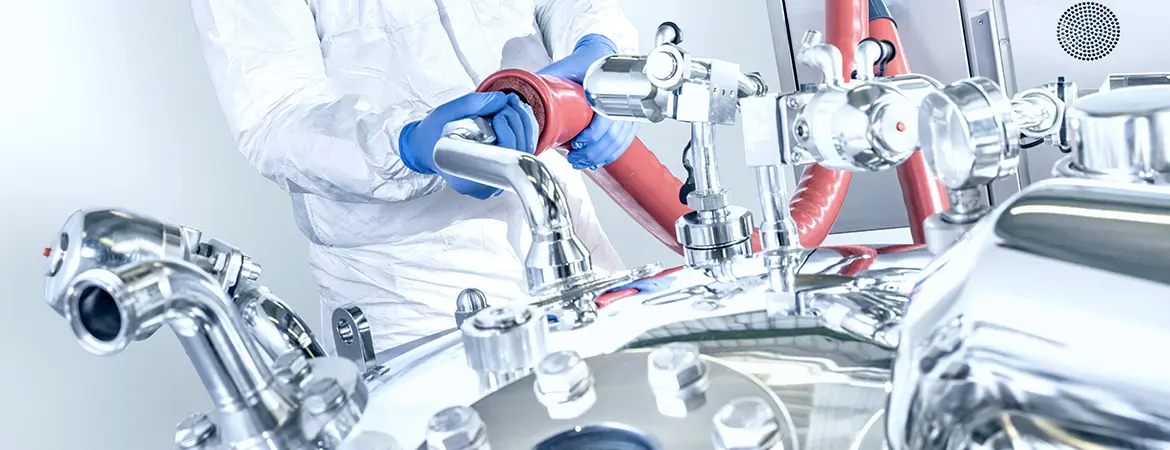 The image shows a person in protective clothing and blue gloves working with shiny metal equipment. They’re handling a red hose connected to pipes and valves. It looks like a clean, controlled setting for manufacturing or lab work.