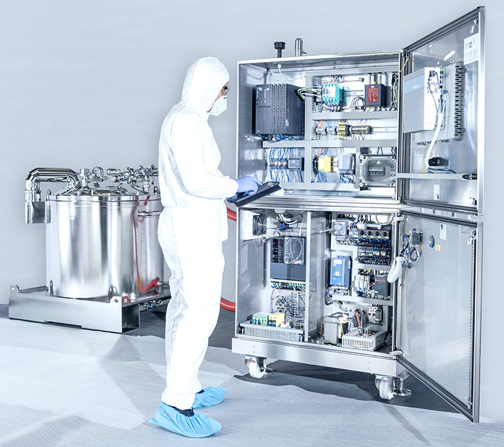 The image shows a person in protective clothing working with industrial equipment. They’re using a tablet near an open control panel full of wires and electronics. Next to them is a large metal tank with pipes and valves.