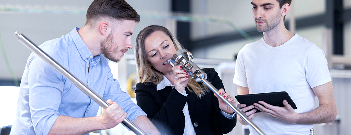 The image shows three people working together in a lab or industrial setting. One is holding a long metal rod, another is inspecting it with a tool, and the third is using a tablet.