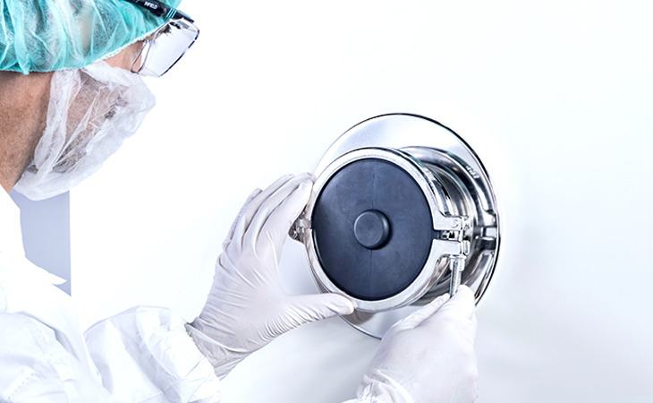 The image shows a person in protective clothing—hair cover, mask, and gloves—working with a round metal device on a white wall.