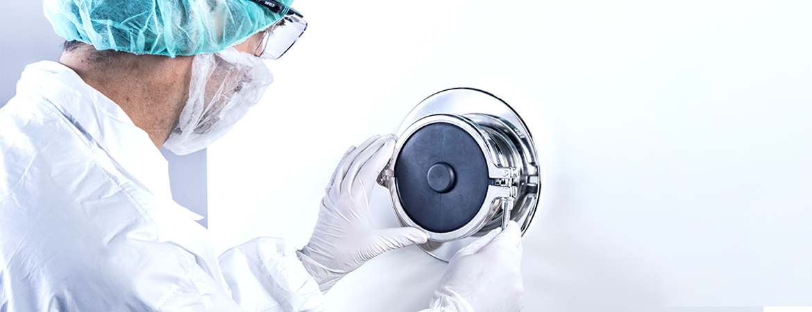 The image shows a person in protective clothing—hair cover, mask, and gloves—working with a round metal device on a white wall.