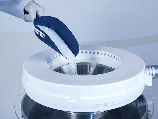 The image shows a person wearing a glove pouring white granules from a blue scoop into a large round device.