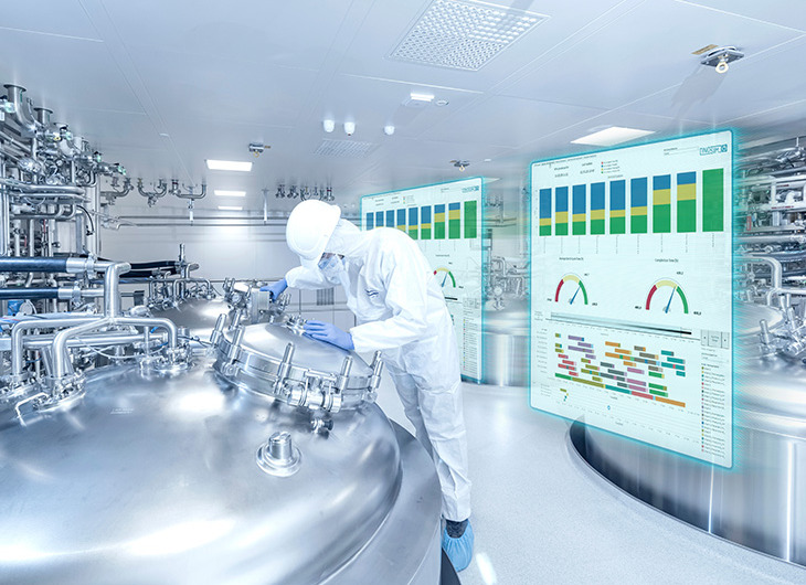 The image shows a person in protective clothing working with large metal equipment in a clean industrial setting. Behind them are two transparent screens showing charts and graphs, suggesting the use of advanced technology to monitor processes.