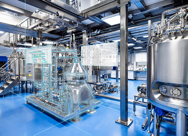 The image shows a clean, modern industrial facility with large metal tanks and complex piping. Some equipment has digital overlays, suggesting the use of augmented reality or smart monitoring. The floor is blue, and the space is brightly lit and well-organized.
