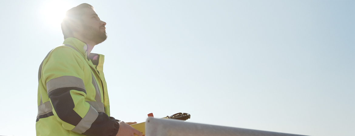 Person wearing a safety jacket outdoors, holding equipment near a metal structure under bright sunlight.