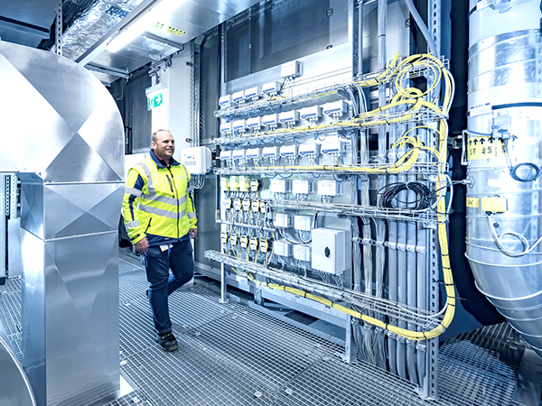The image shows a person in a high-visibility jacket walking through a clean, well-lit industrial facility. The space features organized yellow cables, electrical panels with meters, and metallic surfaces, indicating a controlled and technical environment.