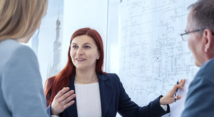 Three people in a professional setting in an engineering discussion, standing near a large technical diagram. One person is pointing at the diagram with a pen while explaining, and the others are attentively observing.