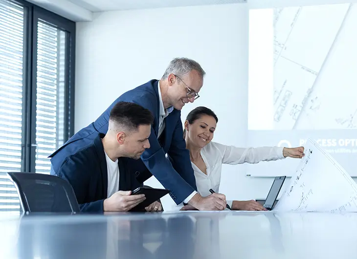 Three people in a meeting room. discussing a plan which is placed on the table in front of them. One person is standing while the other two are sitting.