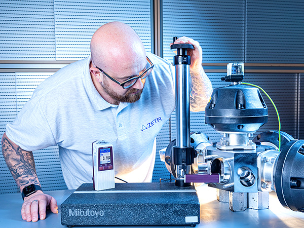 The image shows a person using a precision measuring tool called a height gauge. They’re adjusting it to measure an object, and a digital display nearby shows the value “0.16”. The setting looks like a lab or workshop with metal surfaces and equipment.