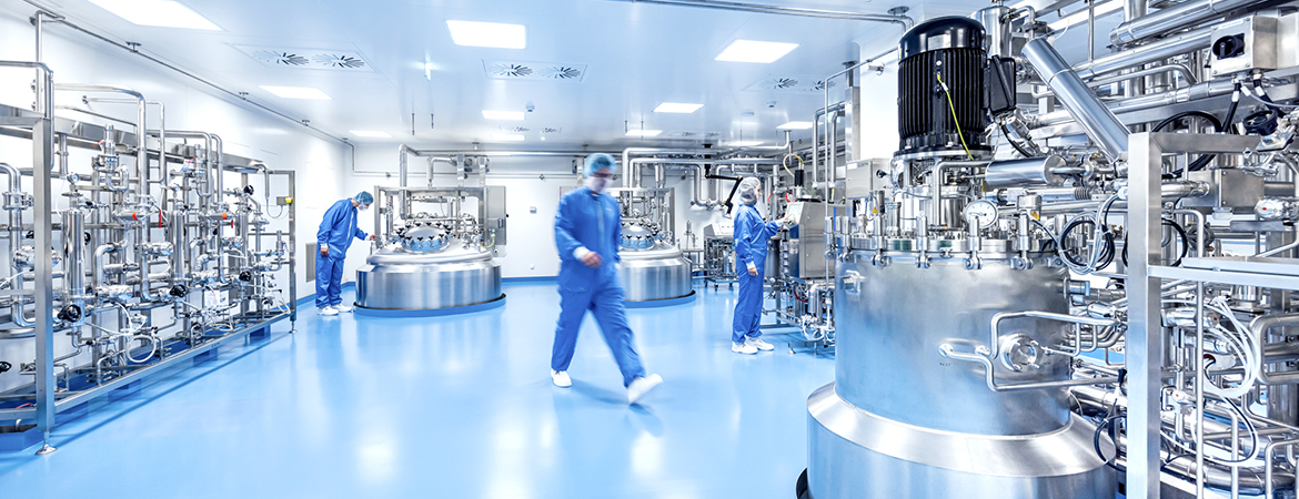 A clean, modern industrial room with shiny metal tanks, pipes, and machines. Three people in blue protective suits and face masks are working.