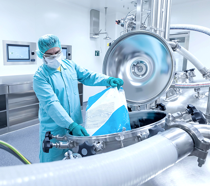 A person in full protective clothing—gowns, gloves, mask, and hair cover—is working in a clean, sterile room, pouring material from a large bag into a metal container.
