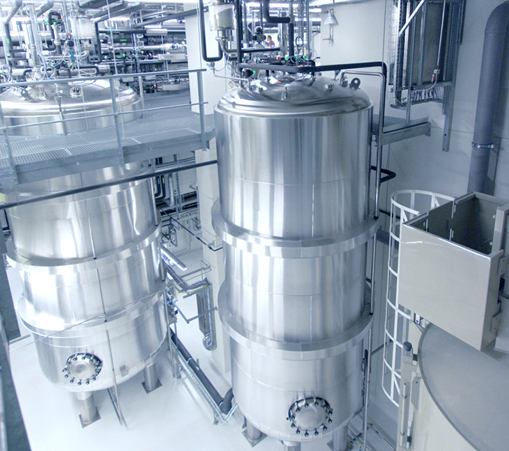 The image shows large stainless steel tanks connected by pipes and valves in a clean industrial facility. A metal walkway above the tanks provides access for maintenance.
