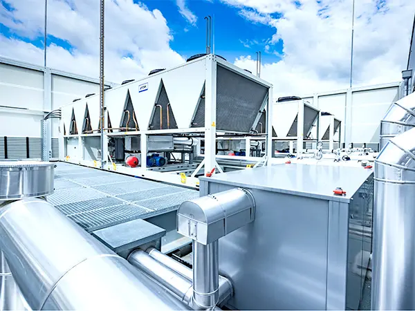 The image shows large air conditioning units on a rooftop. They’re white with triangular vents and connected to pipes and ducts.