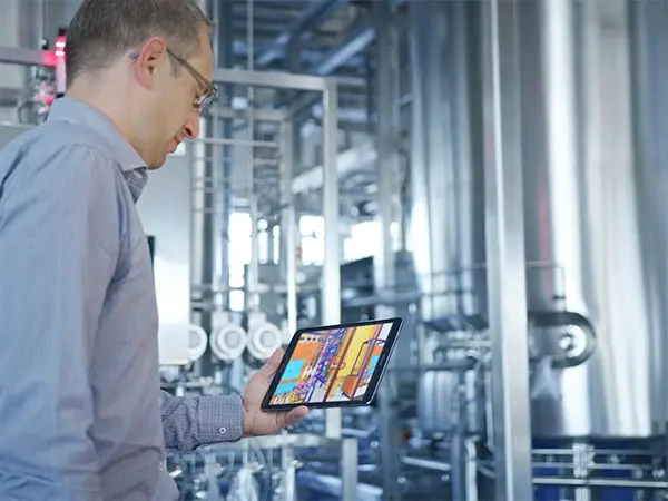 The image shows a person in an industrial setting holding a tablet. The tablet displays a colorful interface, likely used to monitor or control equipment. In the background, there are pipes, tanks, and other machinery.