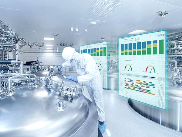 The image shows a person in protective clothing working with large metal equipment in a clean industrial setting. Behind them are two transparent screens showing charts and graphs, suggesting the use of advanced technology to monitor processes.