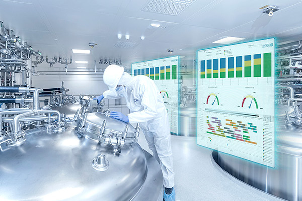 The image shows a person in protective clothing working with large metal equipment in a clean industrial setting. Behind them are two transparent screens showing charts and graphs, suggesting the use of advanced technology to monitor processes.
