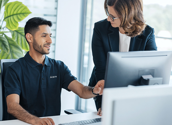 Two people in a modern office setting working together at a desk. One person is seated in front of a computer monitor, wearing a dark polo shirt. The other person is standing beside the desk.