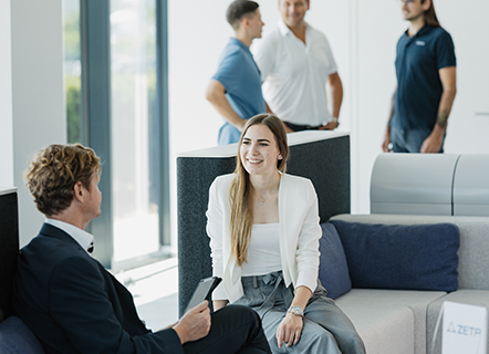 Two people seated on contemporary office lounge furniture having a discussion, while others stand and talk in the background near large windows in a bright, open space.