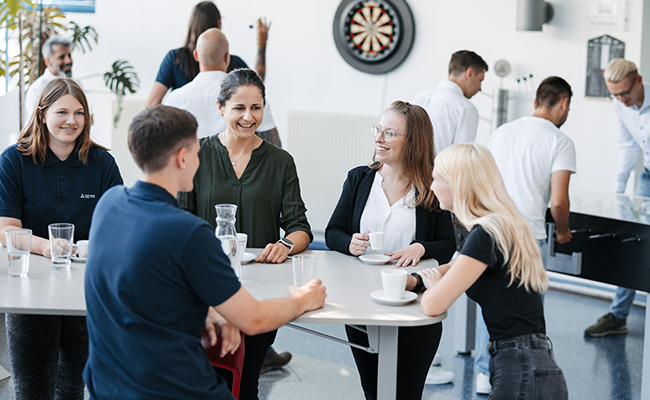 Group of people sitting and standing around a high table with coffee, engaging in conversation in a bright office.
