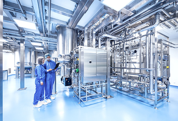 Production area cleanroom with two persons in front of the lower part of a bioreactor that is spanning the ceiling